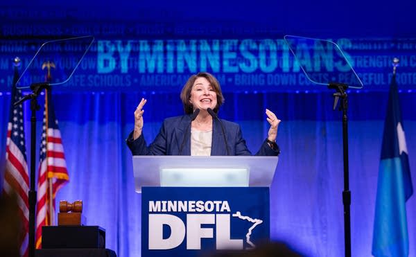 A woman raises her hands while speaking at a podium on stage.