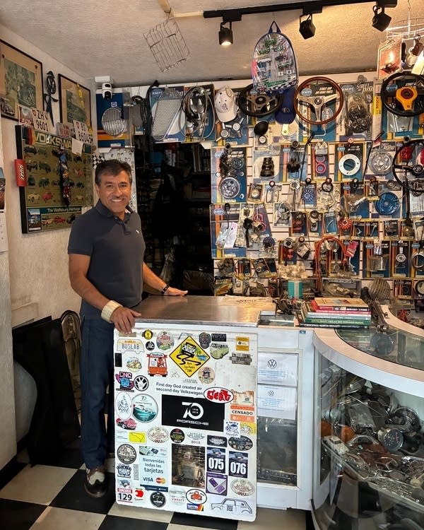 A man stands in a small auto shop surrounded by wheels, art, and stickers
