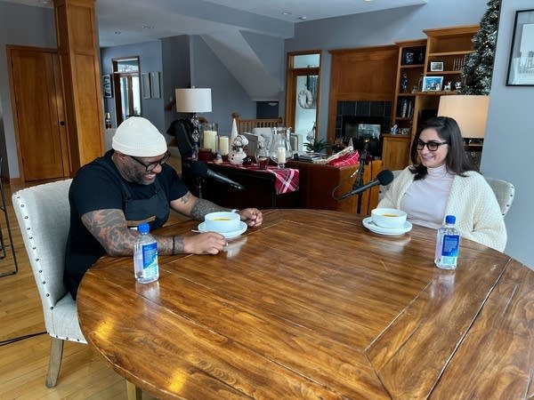 Two people laugh while eating soup at a round wooden table.