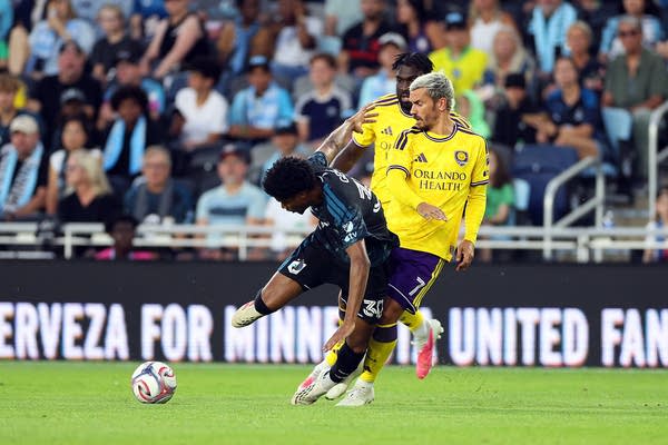 A soccer player in a black uniform trips while being closely chased by two players in yellow uniforms.