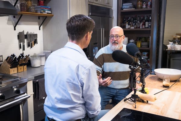 Two people sit in a kitchen with large microphones for an interview.