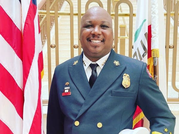 A fire fighter in an official uniform stands next to a U.S. flag.