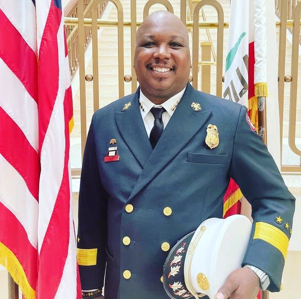 A fire fighter in an official uniform stands next to a U.S. flag.