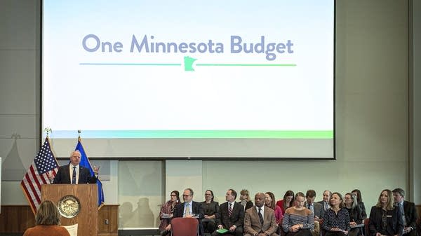 Man stands at podium with others sitting below projection