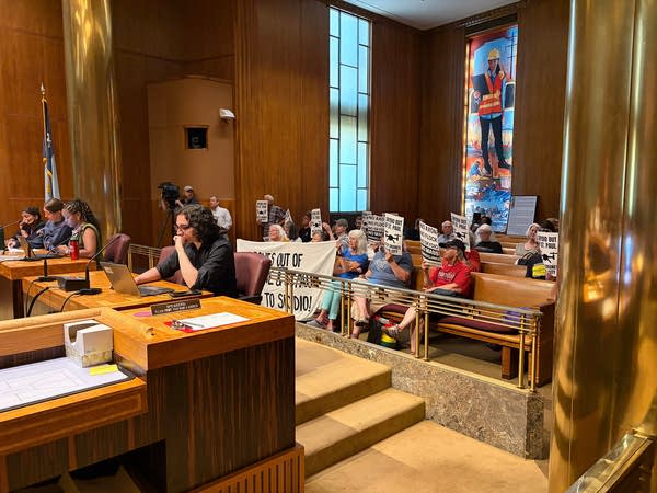 Protesters sit in attendance at a city council meeting while holding signs.