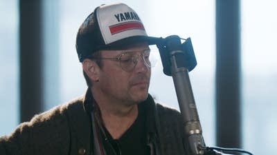 A man in a baseball cap plays guitar in a recording studio