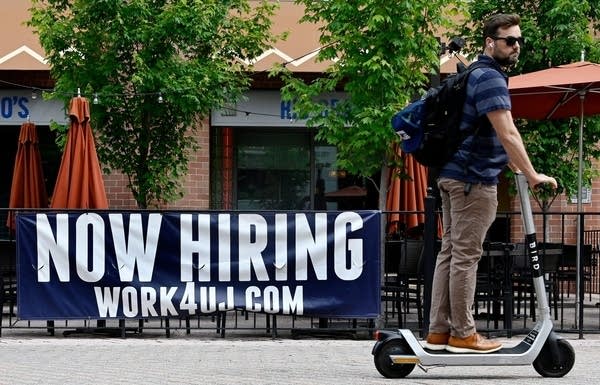 A man riding a scooter passes in front of a "now hiring" sign.