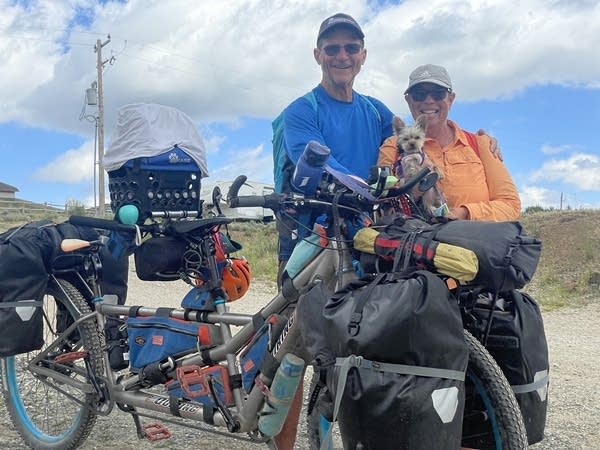 A man in a blue shirt and a woman in an orange jacket hold up a brown Yorkie dog. They are standing behind a tandem bicycle loaded with bags.