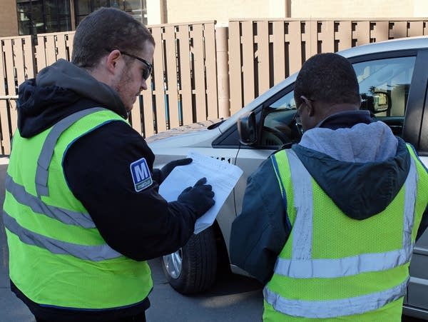 two men stand in a parking lot looking at a list 