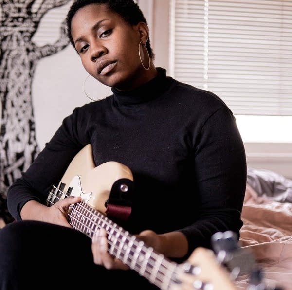 A Black person holds an electric guitar and looks at the camera