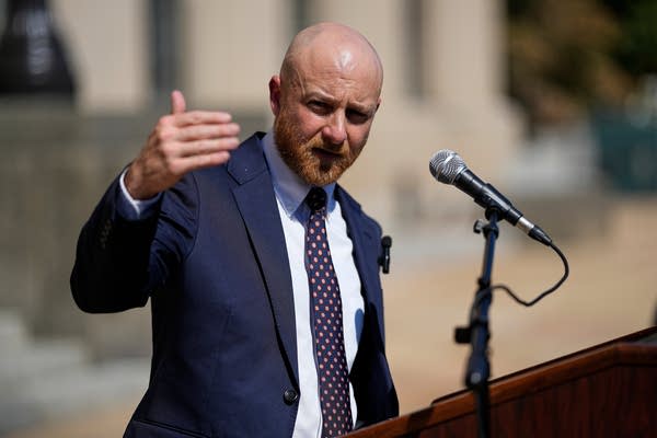 A man in a suit speaks outside at a podium