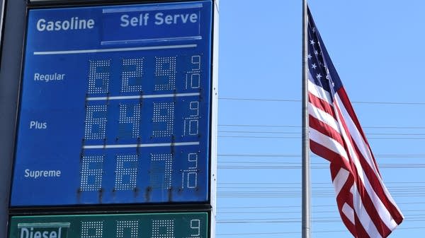 An American flag near a sign displaying gas prices — all north of $6.