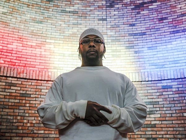 Man standing in white shirt, hands clasped in front of tan brick wall. 