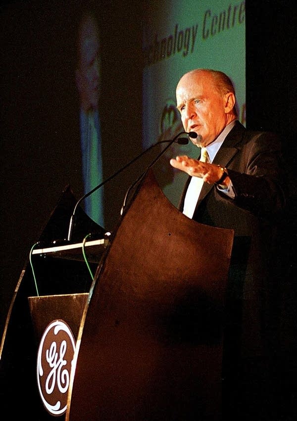 Jack Welch, an older man in a suit, stands at a podium and speaks. The podium bears the GE logo.