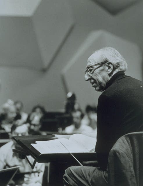 A conductor sits in a chair on the podium during an orchestra rehearsal