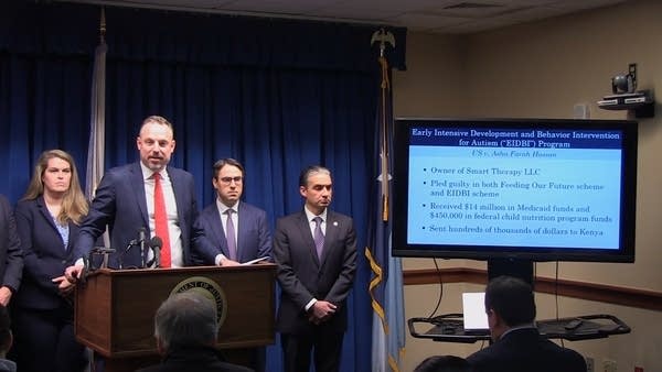 A man stands behind a podium and near a screen displaying a presentation.
