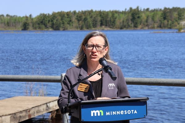 A woman speaking at a podium with a body of water and trees in the background