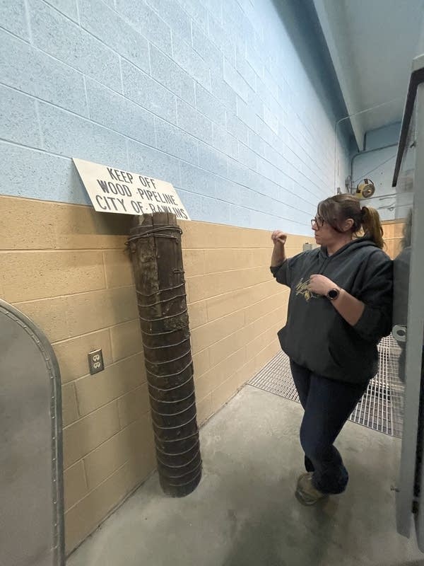 A woman stands in a narrow hallway across from a wooden pipe. A sign above the pipe says " Keep Off Wood Pipeline City of Rawling"