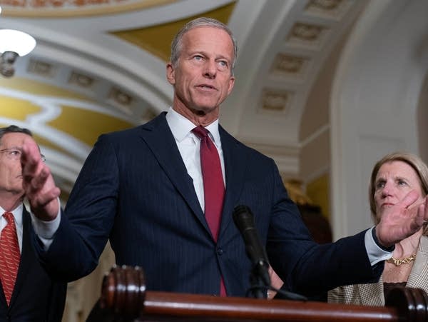A man in a navy suit and red tie raises both his hands at a podium.