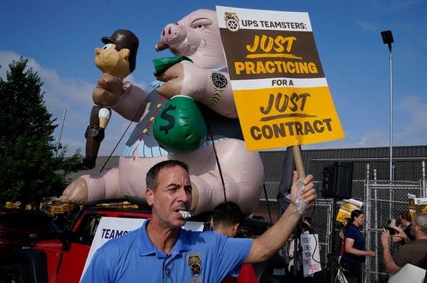 A UPS worker holds up a sign reading "UPS Teamsters: Just practicing for a just contract." In the background, an inflatable pig smoking a cigar sits on top of a red car. The pig holds a UPS worker in one fist and a bag of money in the other.