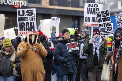 A crowd stands outside holding signs to protest military action in Iran.