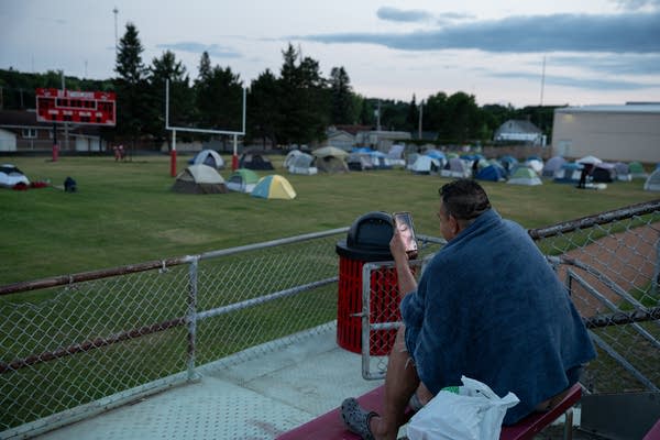 A man looks at his phone while sitting on bleachers with a towel draped over his shoulders. Rows of tents are seen on the field in front of him.