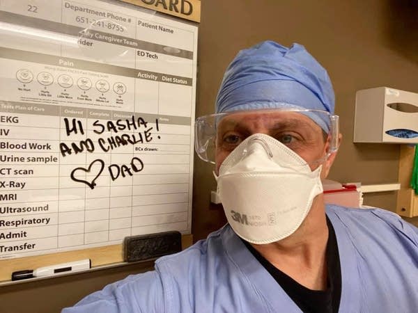 A man wearing scrubs, eye protection and a face mask poses beside a note. 