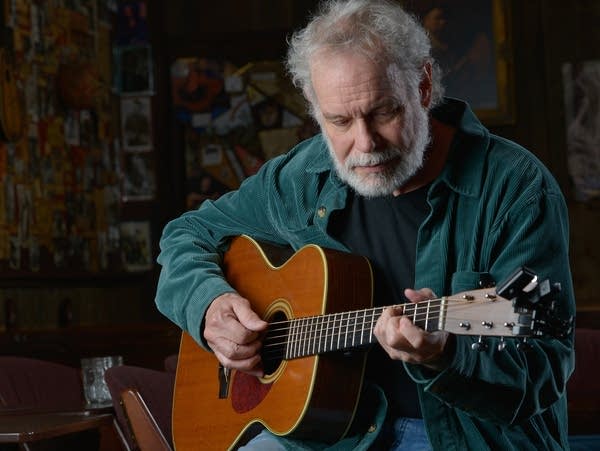 A bearded man sits on a stool and plays guitar