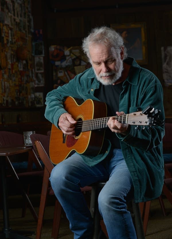 A bearded man sits on a stool and plays guitar