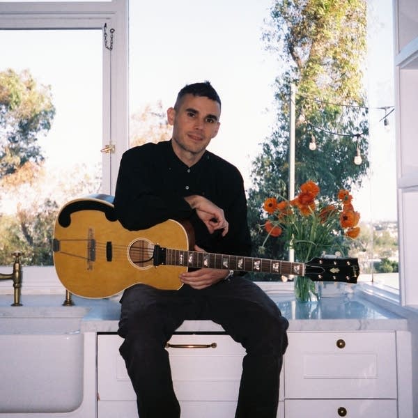 A man with a guitar sits on a kitchen countertop by a large window