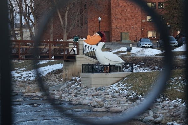 A statue of a pelican near a river. A slight dusting of snow still remains on the ground.
