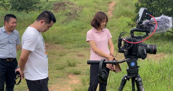 Staffers from the local county propaganda department (seen in pink and white tops) stand in a field. There is a camera in the foreground.