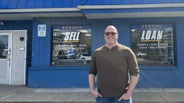 A man stands outside in front of a bright blue building. The building has two windows. One of the windows reads "E & B Coins Sell" and the other reads "E&B Coins Loan."