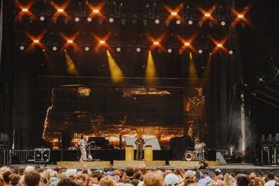 Three musicians in golden masks performing on a large outdoor stage