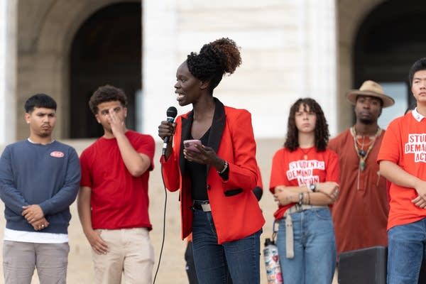 A woman in a red jacket speaks into a microphone with other protesters behind her.