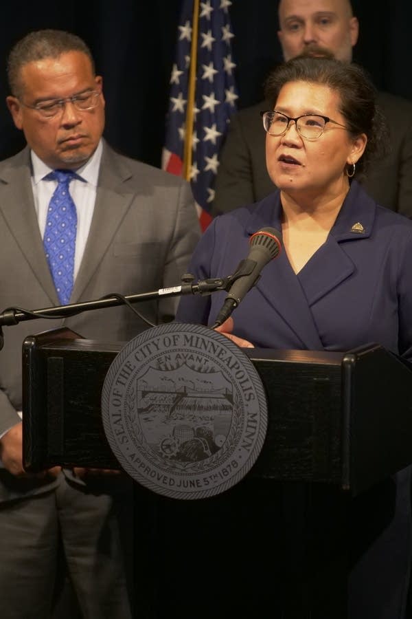 A woman in a navy suit speaks at a podium during a press conference.