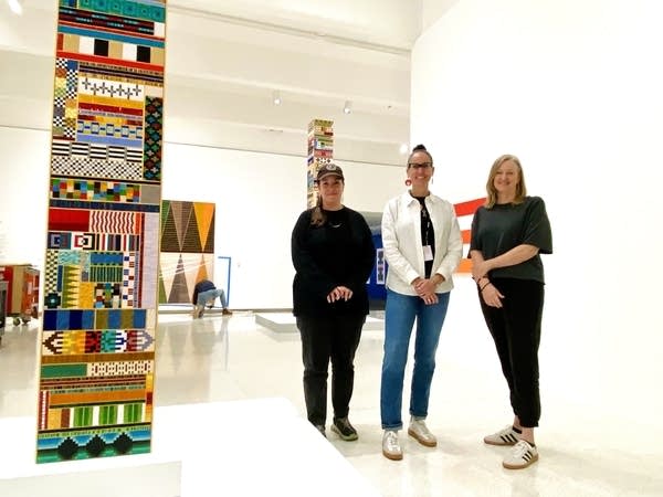 Three women pose next to a multi-colored sculpture in a spacious art gallery.