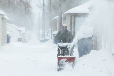 A woman pushes a snow blower down an alley.