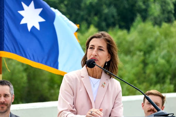 A woman speaks into a microphone. A Minnesota flag blows in the wind behind her.