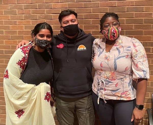 a man and two women stand arm in arm, wearing masks