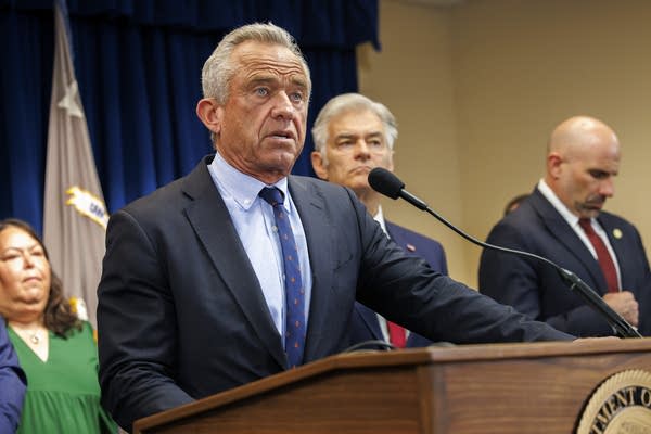 A man in a navy suit and blue shirt speaks into a microphone at a podium.