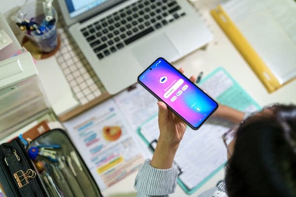 A teenage student interacts with an AI chatbot on a smartphone while studying at a desk with a laptop, notes and stationery. The scene highlights modern learning and technology integration.