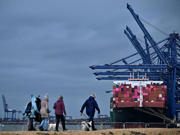 People walk on a beach with a container ship in view.