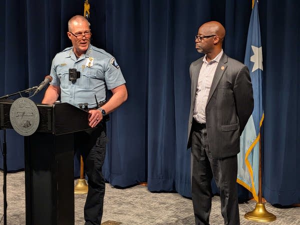 A uniformed police officer speaks at a podium in front of a dark blue curtain while a man in a suit stands next to him.