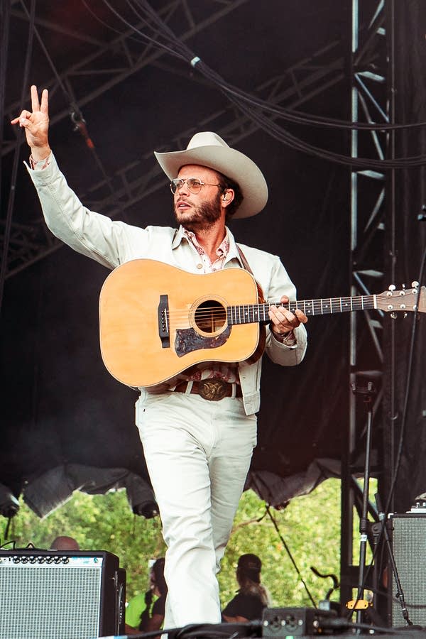 A musician with a guitar walks onstage and greets the audience