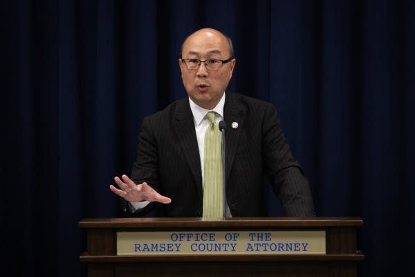 A man in a suit speaks into a microphone at a podium.
