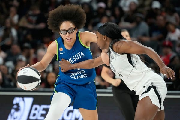 A basketball player in a blue uniform dribbles the ball next to an opposing player in a white uniform.