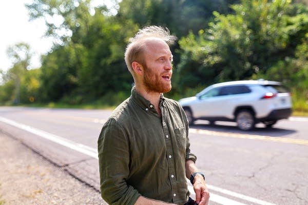 A man stands near the side of a highway with a car passing in the distance.