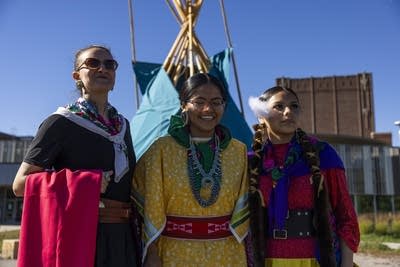 Three dancers dressed in traditional outfits pose in front of a tipi.