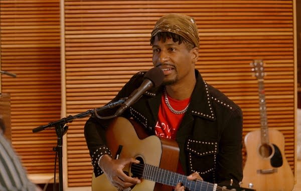 A man speaks into a microphone while holding a guitar